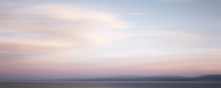 A streaked, periwinkle sky and stratus clouds lit pink and light orange by the setting sun presses down on a strip of sienna brown land and …