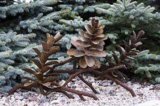 A composition of three small, open pine cones in steel on a thick steel branches sits on the ground as though having just fallen from a tree…