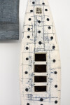 A ceramic canoe shape perforated and marked emerges from a plinth next to a tapestry of blue and light brown that repeats the vertical canoe… Image 4
