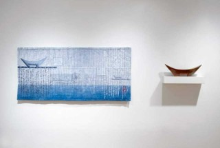 A canoe shaped ceramic vessel glazed in red brown is perched on a floating shelf next to a hanging blue and grey tapestry.