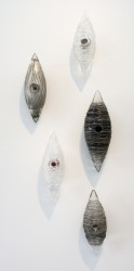 Five blown glass cocoon shapes in clear, charcoal grey, black and silver are curated vertically on the wall in this sculptural installation.