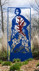 A rectangular aluminum frame planted in the ground contains the cut out figure of a woman in a long dress designed with arabesques, bird, vi…