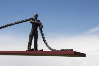 A bronze cast figurine of a man in snowshoes at one end of a red steel plank pulls on a bronze rope that is attached to the other end of the…