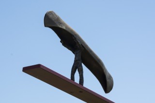 A bronze cast figurine of a man carrying a canoe walks towards the end of a tilted red steel plank.