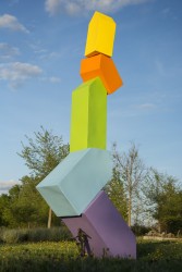 A toy sized man in a winter vest props up a stack of colorful house-shaped steel blocks in this outdoor sculpture by P.