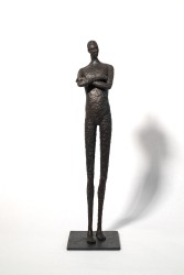He stands tall—a slender naked figure, arms crossed, head slightly tilted.