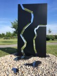 A rectangular, steel outdoor sculpture in black is divided in three parts, like a puzzle, the edges polished and reflective. Image 4
