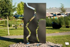 A rectangular, steel outdoor sculpture in black is divided in three parts, like a puzzle, the edges polished and reflective. Image 2