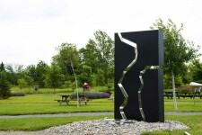 A rectangular, steel outdoor sculpture in black is divided in three parts, like a puzzle, the edges polished and reflective. Image 3