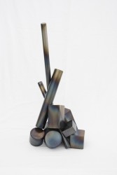 Building blocks of hollow, patinated steel -- cylinders and bars -- are assembled into a tall composition.