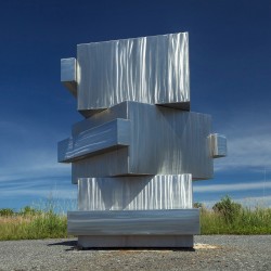 Three large hollow blocks of brushed, reflective steel sided with lengths of steel are stacked at angles.