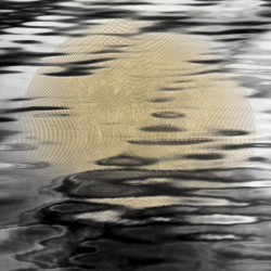 From beneath the ripples on the surface of calm water, a gold sun emerges through precise, geometric perforations.