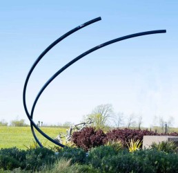 Two narrow steel rods in deep blue rise from the ground and arc towards the sky in this elegant outdoor sculpture by Canadian artist, Shayne…