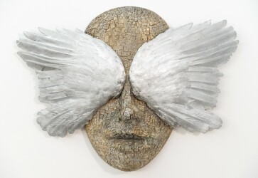 This contemporary wall sculpture is a mask forged from metal adorned with the wings of a bird.