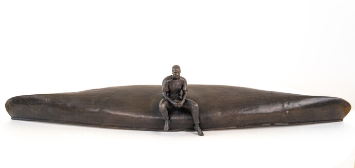 This indoor table top bronze sculpture features a man sitting on an overturned canoe.