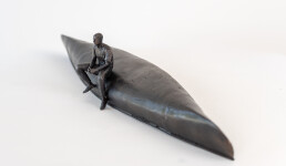 This indoor table top bronze sculpture features a man sitting on an overturned canoe. Image 2
