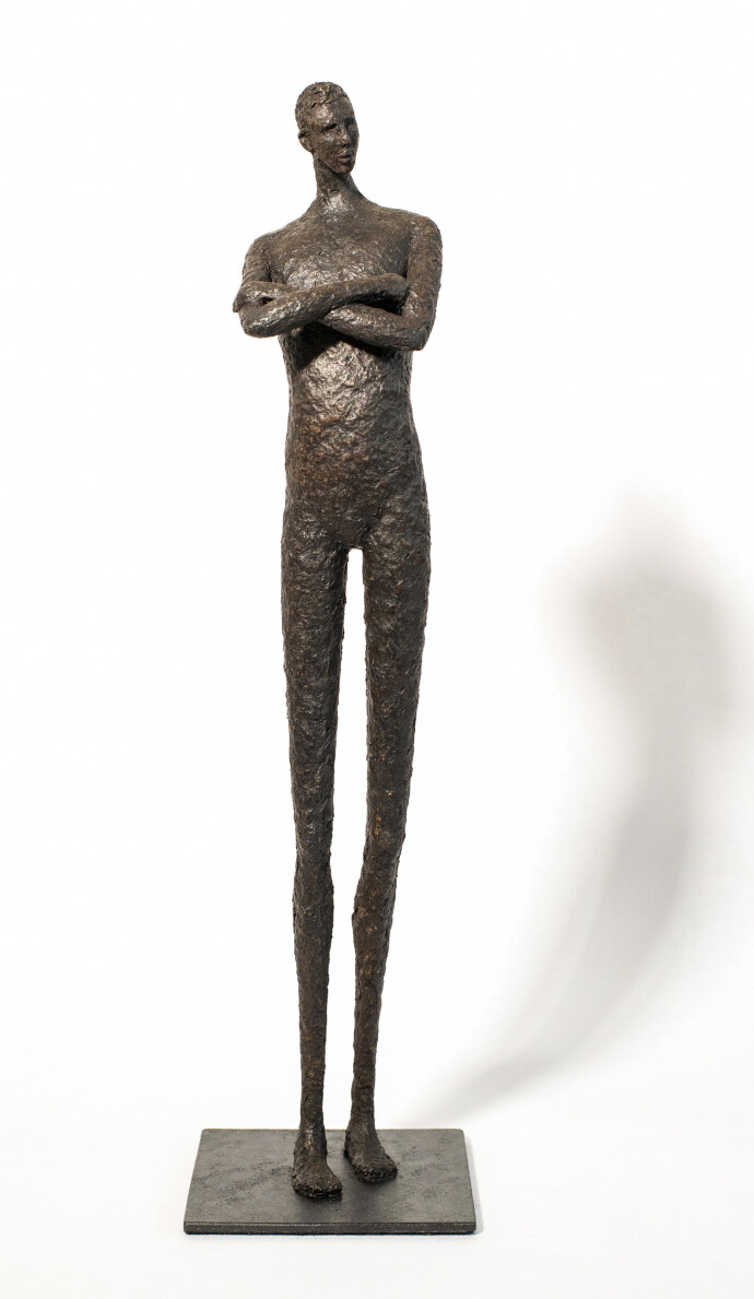 He stands tall—a slender naked figure, arms crossed, head slightly tilted.